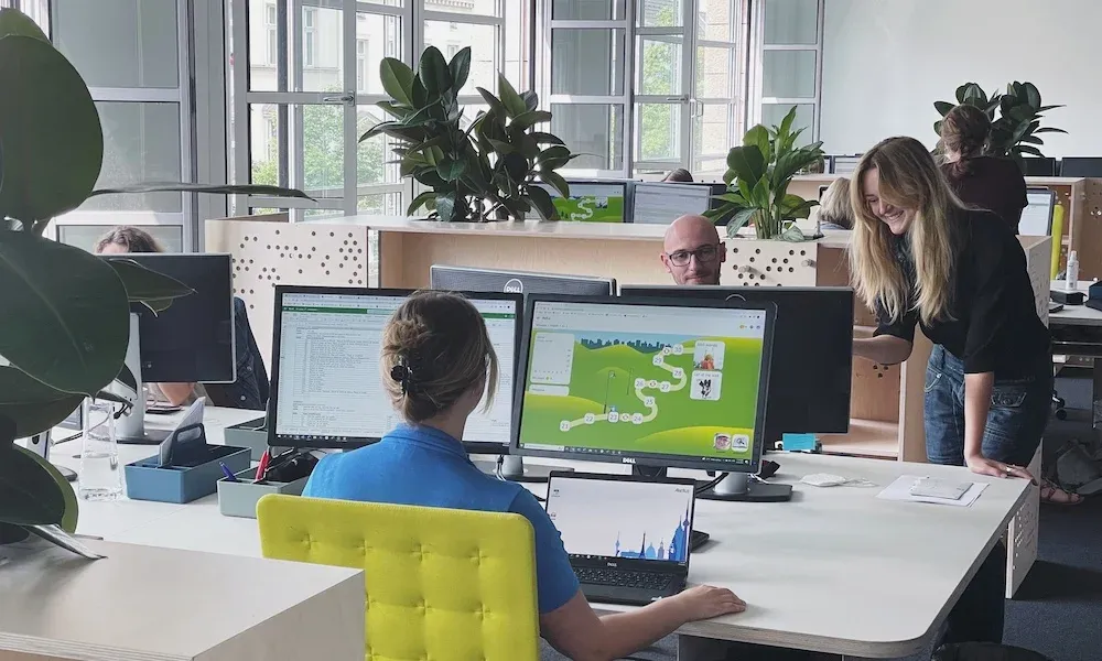 A girl is seated at a desk in a modern office of Akelius Languages, working on developing the Akelius Languages learning platform. She is focused on two monitors displaying the levels of a learning course and design elements. The office environment is neat and professional.